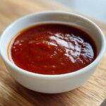 Close-up of rich, dark red homemade bbq sauce served in a small white bowl on a wooden cutting board.