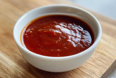 Close-up of rich, dark red homemade bbq sauce served in a small white bowl on a wooden cutting board.