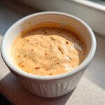 Close-up of creamy, orange-hued big mac sauce speckled with red pepper flakes in a small white bowl.