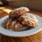 Three soft, golden brown iced oatmeal cookies stacked on a small white plate with vanilla glaze drizzle.
