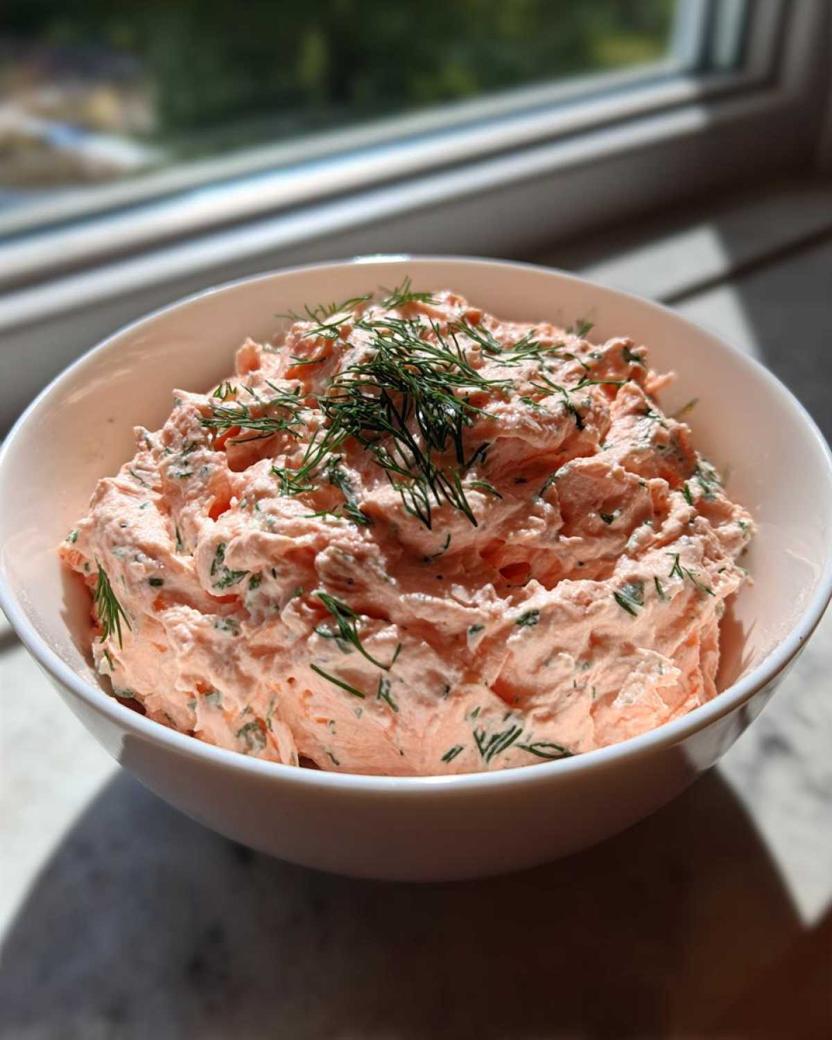 A white bowl filled with creamy, pink smoked salmon dip topped generously with fresh dill.