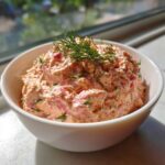 A close-up of creamy, pink smoked salmon dip garnished with fresh dill in a white bowl.