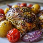 Close-up of a perfectly roasted piece of greek chicken, seasoned with herbs, served with roasted potatoes, red onions, and tomatoes.