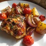 Close-up of a perfectly cooked, glazed greek chicken thigh served with roasted potatoes, red onions, and cherry tomatoes.
