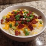 A close-up of a bowl of creamy loaded baked potato soup topped generously with shredded cheddar cheese, crispy bacon bits, and sliced green onions.