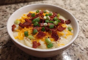 A close-up of a bowl of creamy loaded baked potato soup topped generously with shredded cheddar cheese, crispy bacon bits, and sliced green onions.