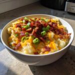 A close-up of a white bowl filled with creamy loaded baked potato soup, topped generously with shredded cheddar, crispy bacon bits, and fresh green onions.