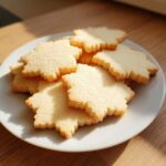 A stack of freshly baked, golden maple leaf shaped sugar cookie treats resting on a white plate.
