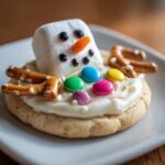 A close-up of a single melted snowman cookies featuring a marshmallow head, pretzel arms, and colorful candy buttons.