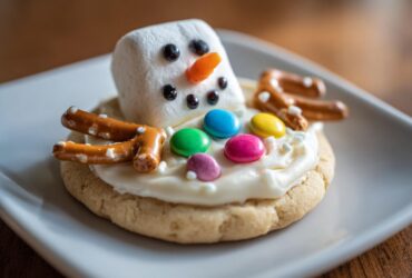 A close-up of a single melted snowman cookies featuring a marshmallow head, pretzel arms, and colorful candy buttons.