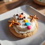 A single melted snowman cookie decorated with white frosting, pretzel arms, candy buttons, and chocolate chip eyes.