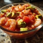 Close-up of a glass bowl filled with vibrant Mexican shrimp cocktail, featuring shrimp, tomatoes, and chunks of avocado.