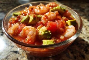 Close-up of a glass bowl filled with vibrant Mexican shrimp cocktail, featuring shrimp, tomatoes, and chunks of avocado.