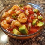 Close-up of a glass bowl filled with vibrant mexican shrimp cocktail, featuring shrimp, avocado chunks, and tomato sauce.