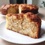 Close-up of a moist slice of Jewish apple cake showing tender crumb and baked apple pieces, with the rest of the cake blurred in the background.
