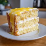 Close-up of a moist slice of two-layer lemon cake recipe with white frosting and a bright yellow lemon drizzle.