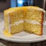 Close-up of a moist slice of lemon cake recipe, showing two layers filled with cream and topped with dripping lemon curd.