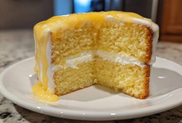 Close-up of a moist slice of lemon cake recipe, showing two layers filled with cream and topped with dripping lemon curd.