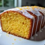 A close-up of a freshly baked lemon loaf sliced open, topped with white glaze and orange zest.