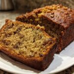 A close-up of a freshly baked, moist loaf of sugar free banana bread, partially sliced and topped with walnuts.