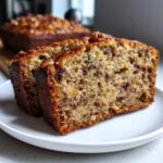 Two thick slices of moist sugar free banana bread, showing nuts and dark banana flecks, served on a white plate.