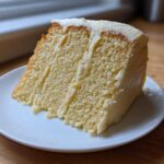 A generous slice of moist vanilla cake recipe layered with creamy white frosting, sitting on a small white plate.