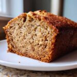 Close-up of a thick slice of moist vegan banana bread showing its crumb texture, resting on a white plate.
