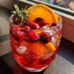 Close-up of a glass filled with bright red non-alcoholic christmas punch, garnished with fresh cranberries, an orange slice, and rosemary.
