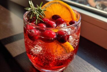 Close-up of a glass filled with bright red non-alcoholic christmas punch, garnished with fresh cranberries, an orange slice, and rosemary.