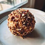 Close-up of a homemade pineapple cheese ball coated entirely in chopped pecans, sitting on a light plate.