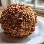 A close-up of a perfectly formed pineapple cheese ball completely coated in chopped pecans, resting on a white plate.
