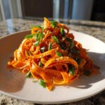 A close-up of a vibrant raw carrot salad made with shaved carrot ribbons, topped with sesame seeds and fresh green onions.