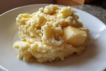 A close-up of fluffy roasted garlic mashed potatoes topped with melted butter and herbs on a white plate.