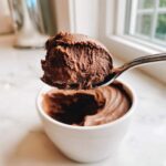 A spoonful of rich, airy chocolate mousse being lifted above a white bowl of the dessert.