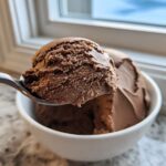 A spoonful of rich, dark chocolate mousse being lifted from a white bowl near a window.