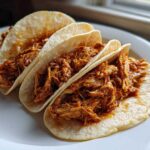 Three soft corn tortillas filled generously with saucy shredded chicken tacos, ready to eat.