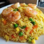 A mound of yellow shrimp fried rice topped with perfectly cooked pink shrimp, peas, and carrots.