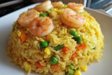 A mound of yellow shrimp fried rice topped with perfectly cooked pink shrimp, peas, and carrots.