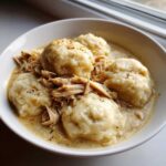 A close-up of a white bowl filled with creamy slow cooker chicken and dumplings, topped with shredded chicken and pepper.