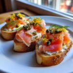 Three slices of crostini topped with cream cheese, smoked salmon, capers, lemon zest, and dill.