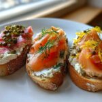 Three slices of crostini topped with cream cheese, smoked salmon, capers, dill, and lemon zest.