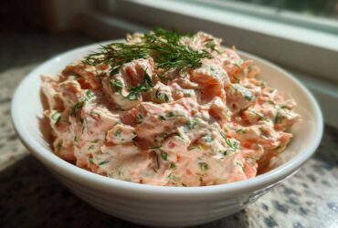 A close-up of a white bowl filled with creamy, chunky smoked salmon dip garnished with fresh dill.