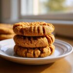 Three soft peanut butter cookies stacked on a white plate, showing classic crisscross fork marks.