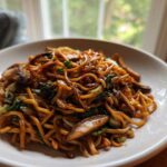 A close-up shot of a white bowl filled with glossy, savory soy sauce fried noodles mixed with mushrooms and green vegetables.