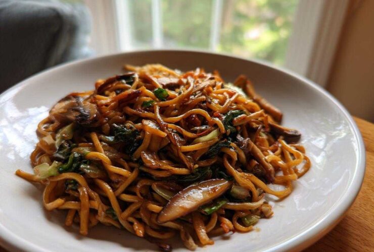A close-up shot of a white bowl filled with glossy, savory soy sauce fried noodles mixed with mushrooms and green vegetables.
