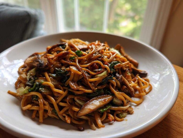 A close-up shot of a white bowl filled with glossy, savory soy sauce fried noodles mixed with mushrooms and green vegetables.
