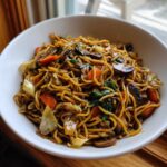 A close-up of a white bowl filled with savory soy sauce fried noodles mixed with mushrooms, carrots, and cabbage.