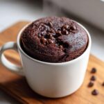 A freshly baked, steaming chocolate chip mug cake rising over the rim of a white ceramic mug.
