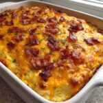 A baked popper dip topped with melted cheddar cheese, crispy bacon bits, and chives in a white rectangular dish.