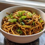 A close-up of rich, saucy garlic noodles topped generously with bright green sliced scallions in a white bowl.